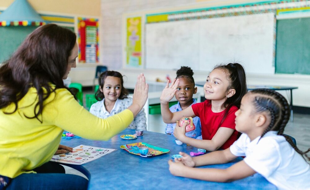 A teacher high-fives a smiling young student in a colorful classroom, surrounded by children engaged in a hands-on learning activity.