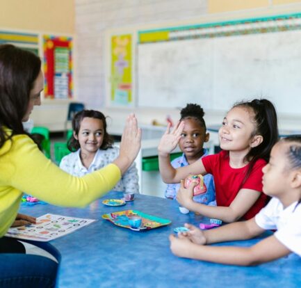 A teacher high-fives a smiling young student in a colorful classroom, surrounded by children engaged in a hands-on learning activity.